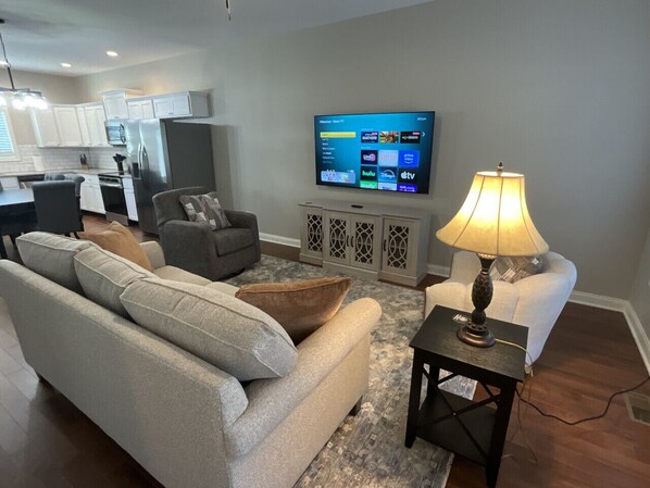 Ein Wohnzimmer mit grauem Sofa, einem weißen Sessel und einem Couchtisch. Ein Flachbildfernseher ist an der Wand montiert und eine Lampe steht auf einem Beistelltisch.