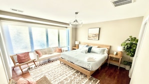 Un dormitorio moderno con una cama grande, una zona de estar con sofá y sillones, una alfombra estampada y un ventanal con persianas.