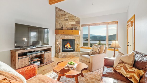 A modern living room with a stone fireplace, a flat-screen TV, and a large window offering a scenic view. - Walk to Town Center Shops & Restaurants, Private Hot Tub, 3BR + Loft (Big Sky)