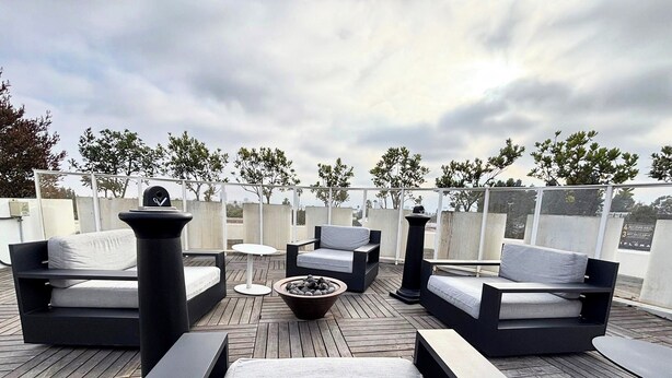 A rooftop terrace with modern outdoor furniture, including sofas and a round table, surrounded by a wooden deck and a view of the city skyline.
