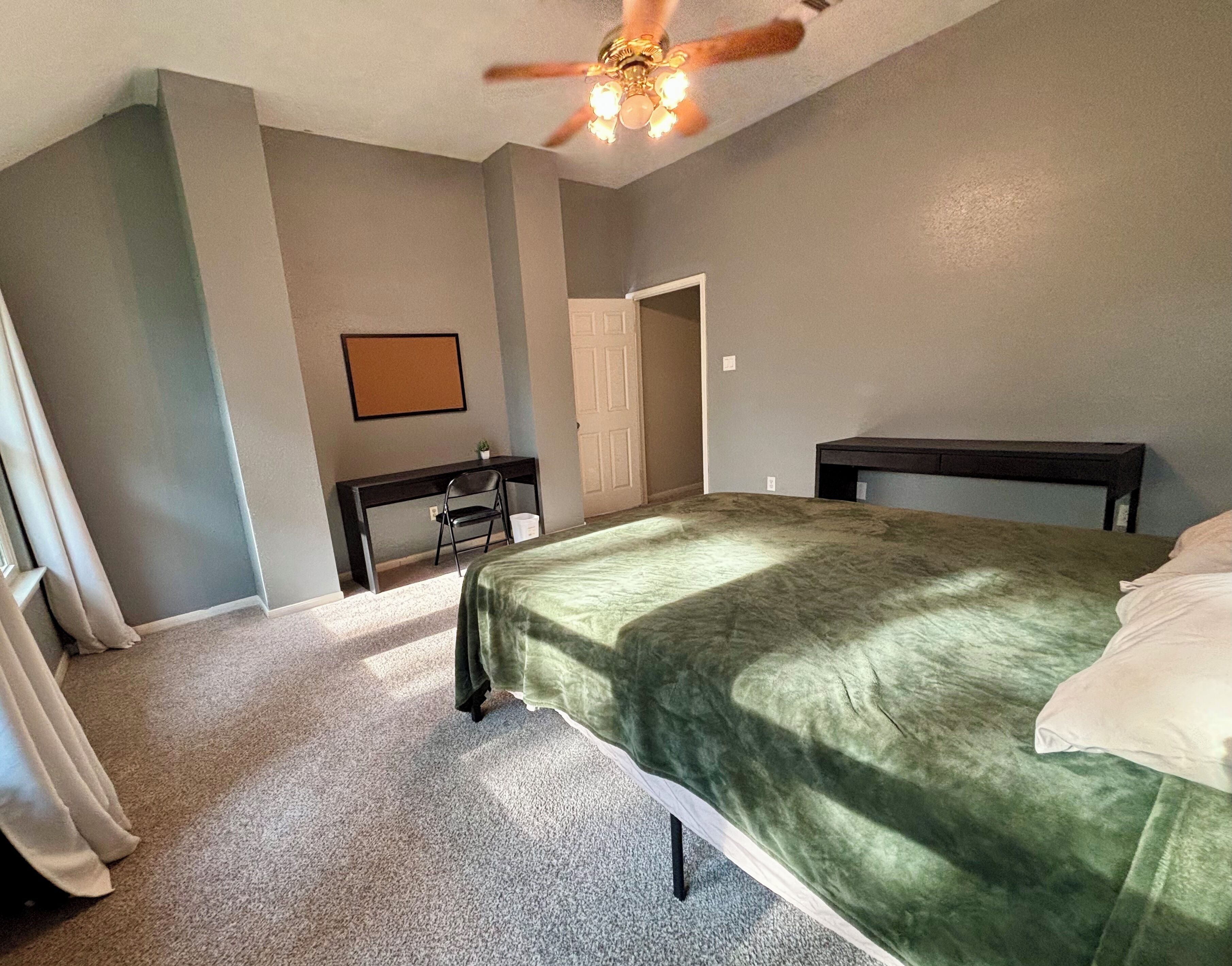 A bedroom with a green bedspread, a desk with a chair, a ceiling fan, and a door leading to another room.