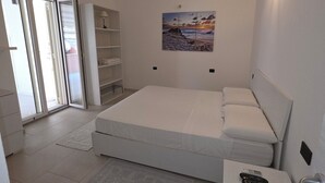 Une chambre moderne avec un lit simple, un petit bureau, une table de chevet, un tableau au mur et une porte-fenêtre coulissante donnant sur un balcon.