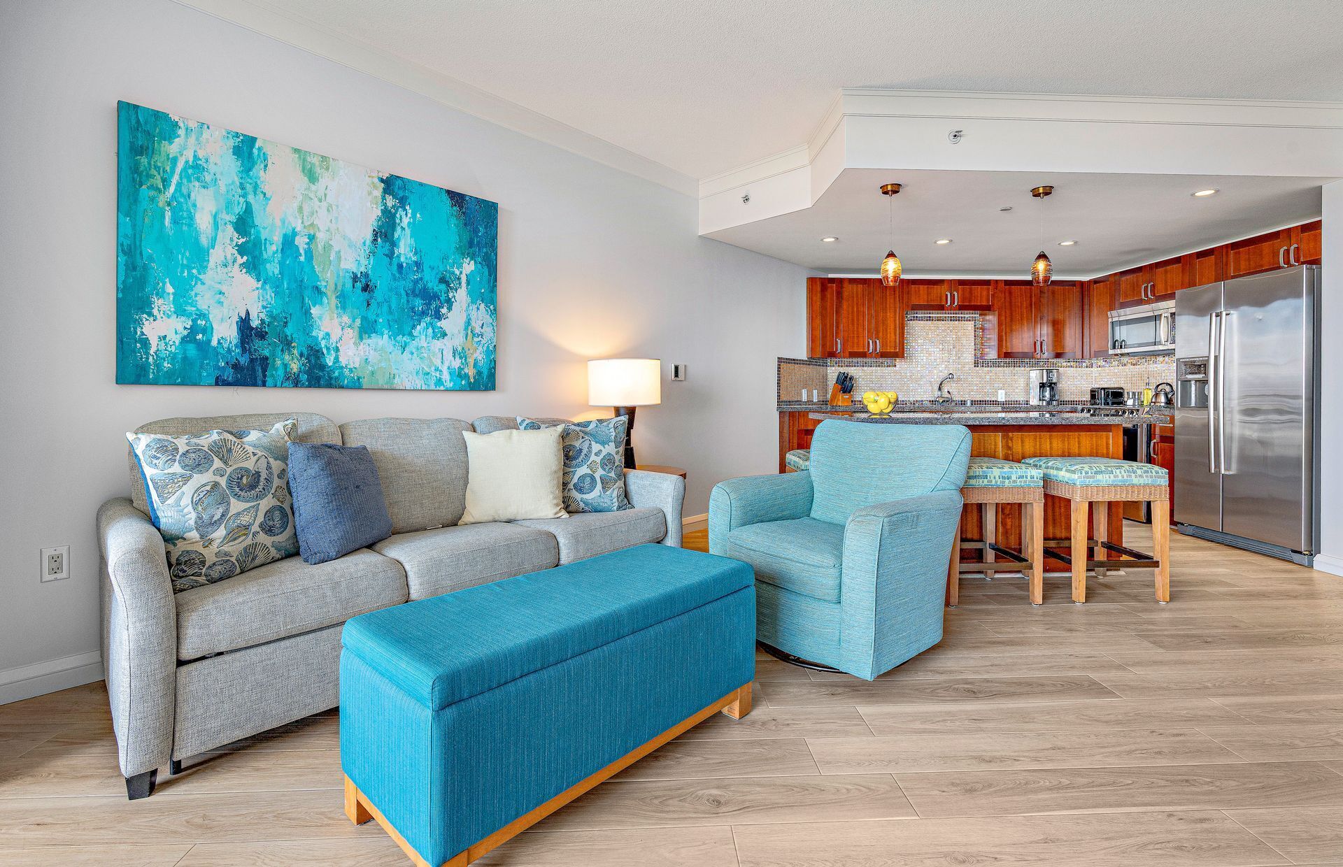Un soggiorno moderno con un divano grigio, un pouf blu e una poltrona, una cucina con mobili in legno ed elettrodomestici in acciaio inossidabile, e un grande quadro astratto appeso al muro.
