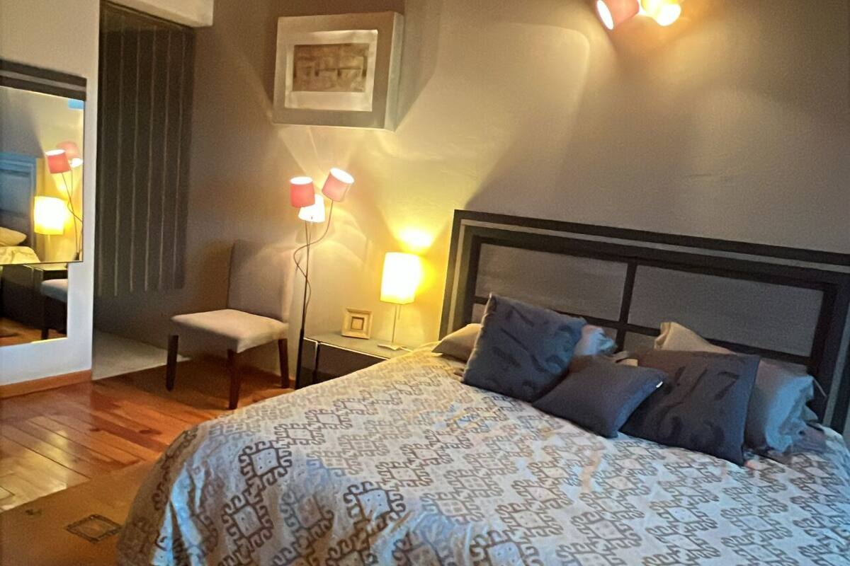 A bedroom with a wooden ceiling, a bed with patterned bedding, two bedside lamps, a chair, and a framed picture on the wall.