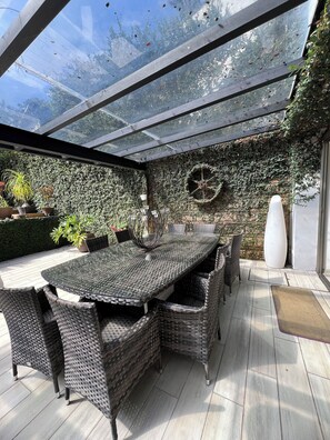 Eine überdachte Terrasse mit Glasdach, Korbmöbeln und verschiedenen Topfpflanzen.
