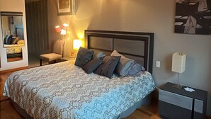 Un dormitorio con techo de madera, una cama con ropa de cama estampada, una mesita de noche, una lámpara de noche, un espejo y un cuadro en la pared.