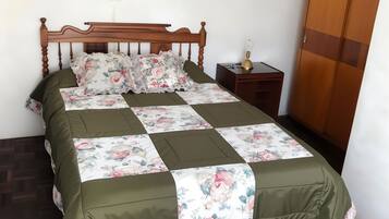 A neatly made bed with a floral bedspread and green curtains, a wooden wardrobe, and a small bedside table with a lamp.