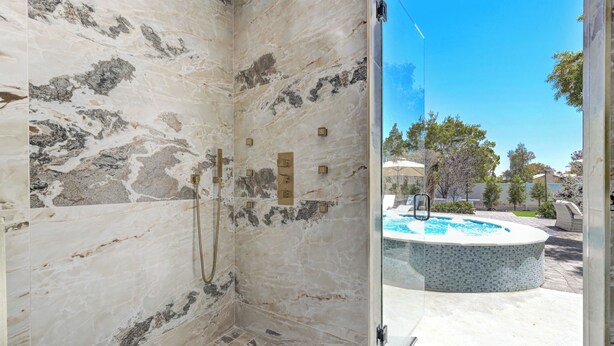 Un baño moderno con mampara de vidrio, paredes de mármol y vistas a un jacuzzi y zona exterior.