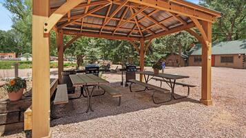 A covered picnic area with wooden benches and tables, surrounded by gravel and trees.