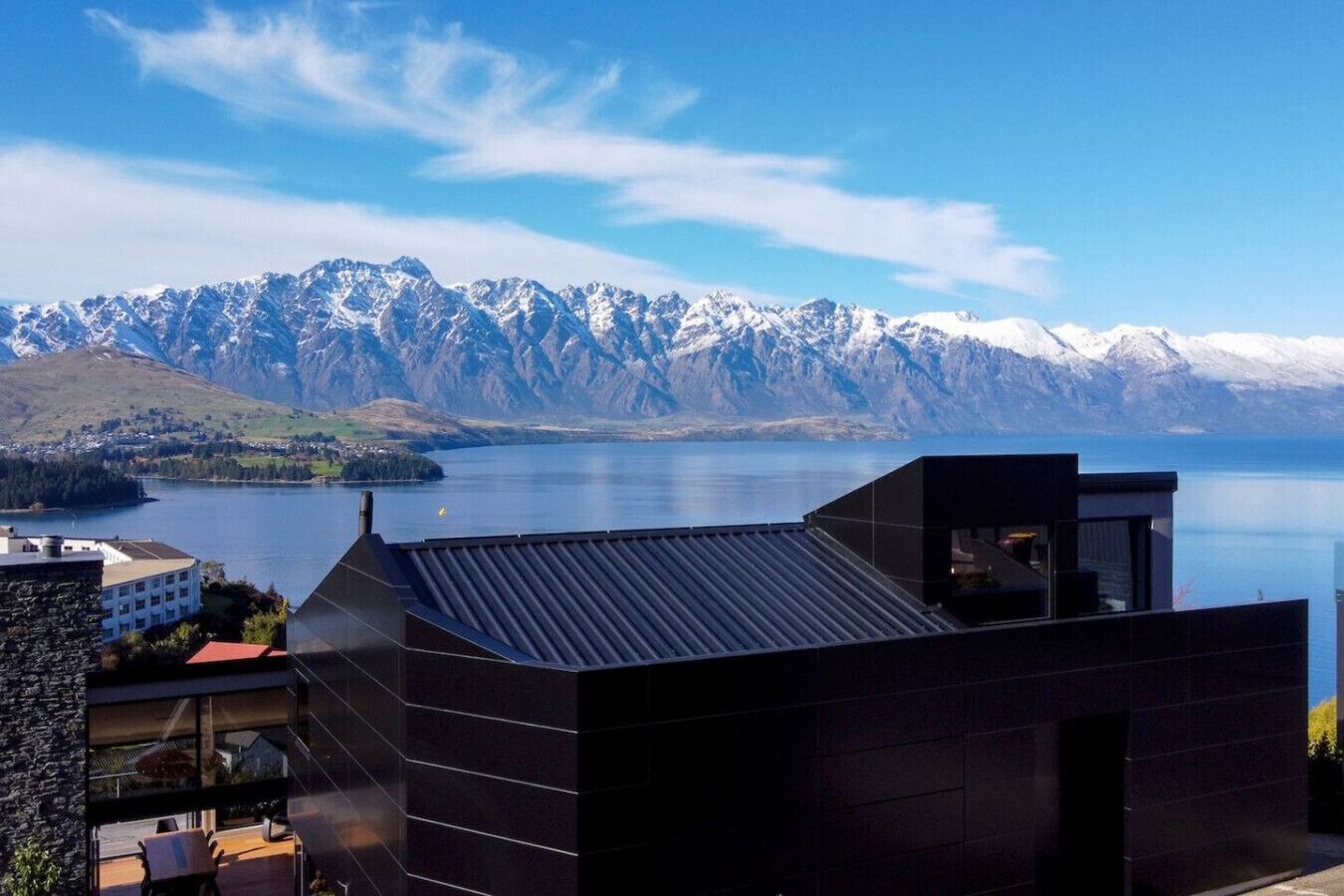 A modern building with a dark roof, overlooking a lake and snow-capped mountains.