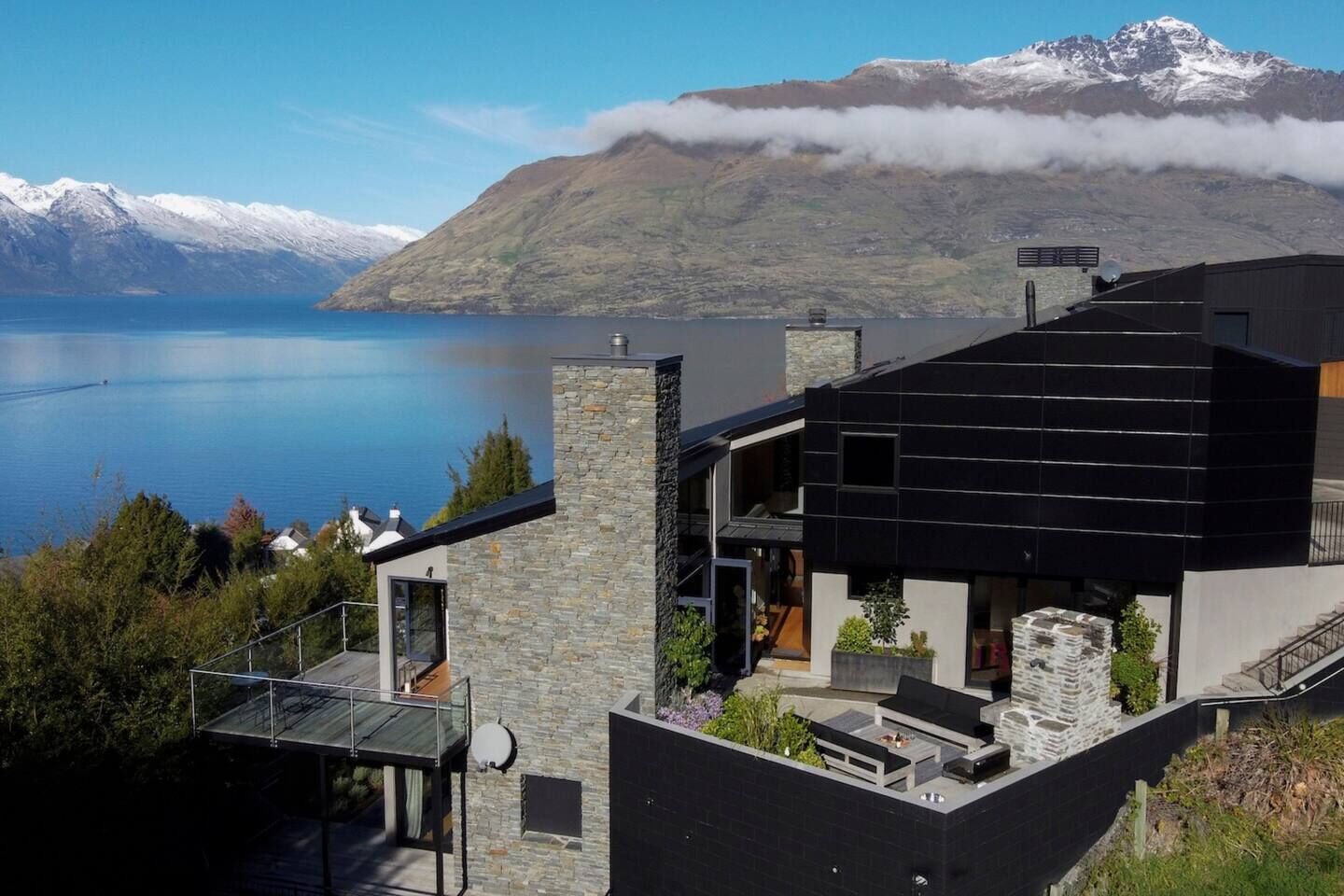 Una casa moderna con fachada de piedra, techo negro y vista a un lago y montañas.