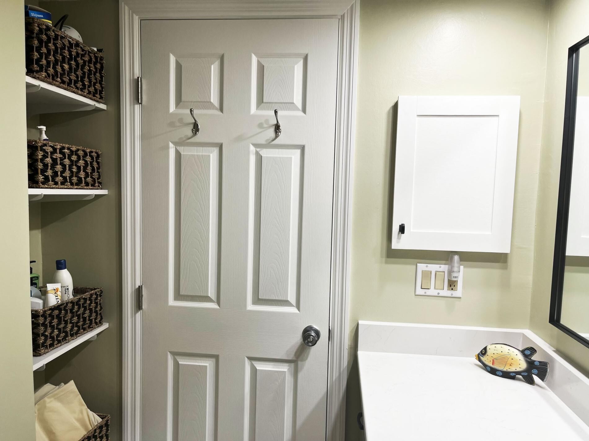 A bathroom with a white door, a white sink, and a fish-shaped soap dish. There are shelves with baskets and toiletries.