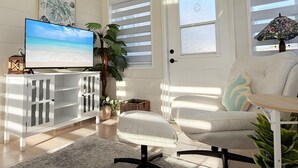 A living room with a television displaying a beach scene, a white cabinet, a white chair, and a door with windows. - Coastal Chic 2BR – Modern Stay Near Manasota Key (Englewood)