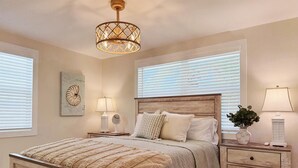 A bedroom with a wooden bed, a nightstand with a lamp, a bedside table with a mirror, and a wall-mounted light fixture. - Coastal Chic 2BR – Modern Stay Near Manasota Key (Englewood)