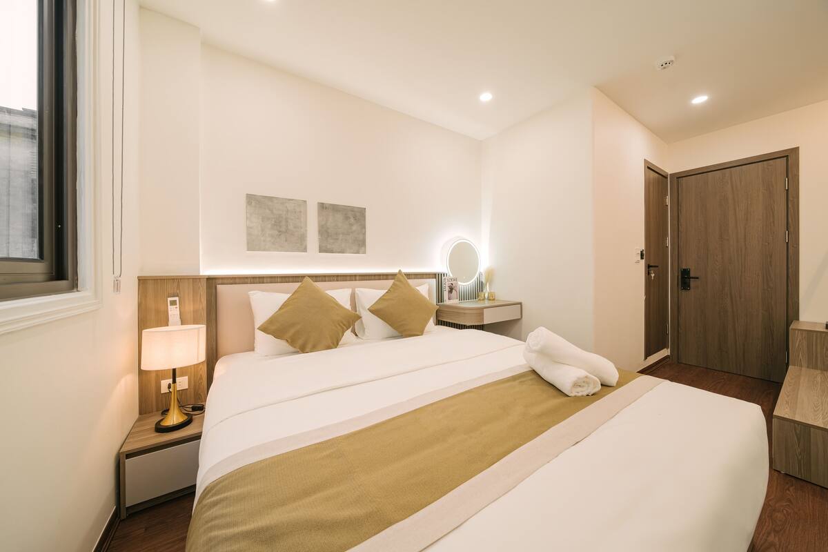 Habitación de hotel con una cama grande, una mesita de noche con lámpara, un espejo y un armario de madera.
