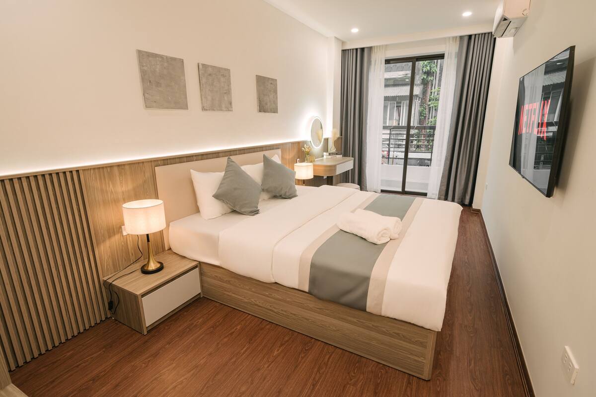 Habitación de hotel con cama, lámpara de noche, una mesita con espejo, televisor y ventana con cortinas.