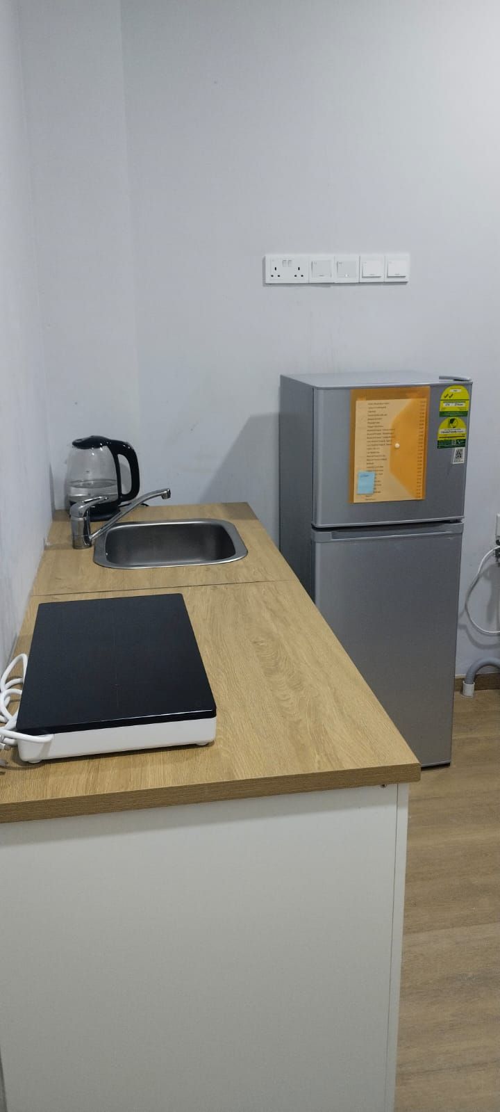 Una kitchenette pequeña con un lavamanos, una tetera, un refrigerador y un calentador portátil sobre una encimera de madera.