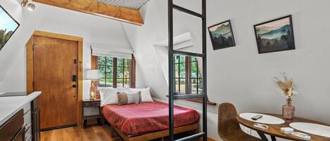 A compact bedroom with a bed, bedside table, lamp, and a window with a view of trees.