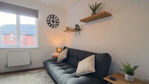 A living room with a grey sofa, two white pillows, a wooden side table with a plant, a wall clock, and a radiator. - Derby Luxury Apartment - Foss Road- Sleeps4 (Derby)