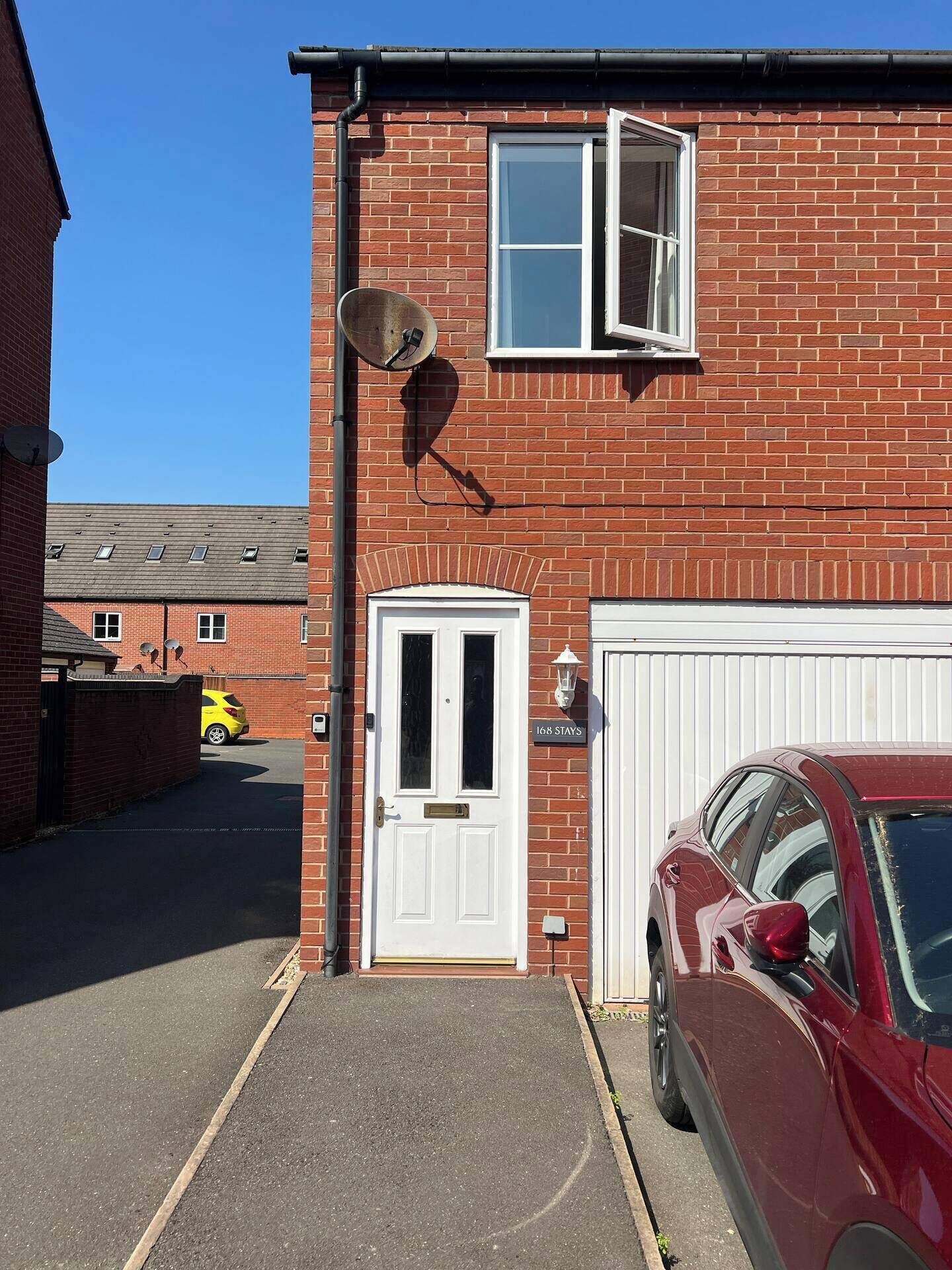 Un bâtiment en briques rouges avec une porte blanche et un garage. Une antenne parabolique est fixée au mur, et une voiture est garée dans l’allée.