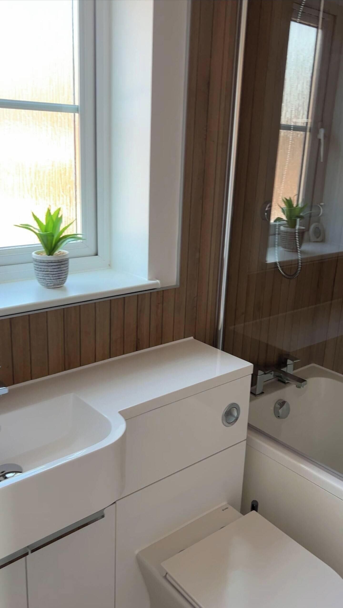 A bathroom with a white sink, a mirror, a shower, and a window with a plant.