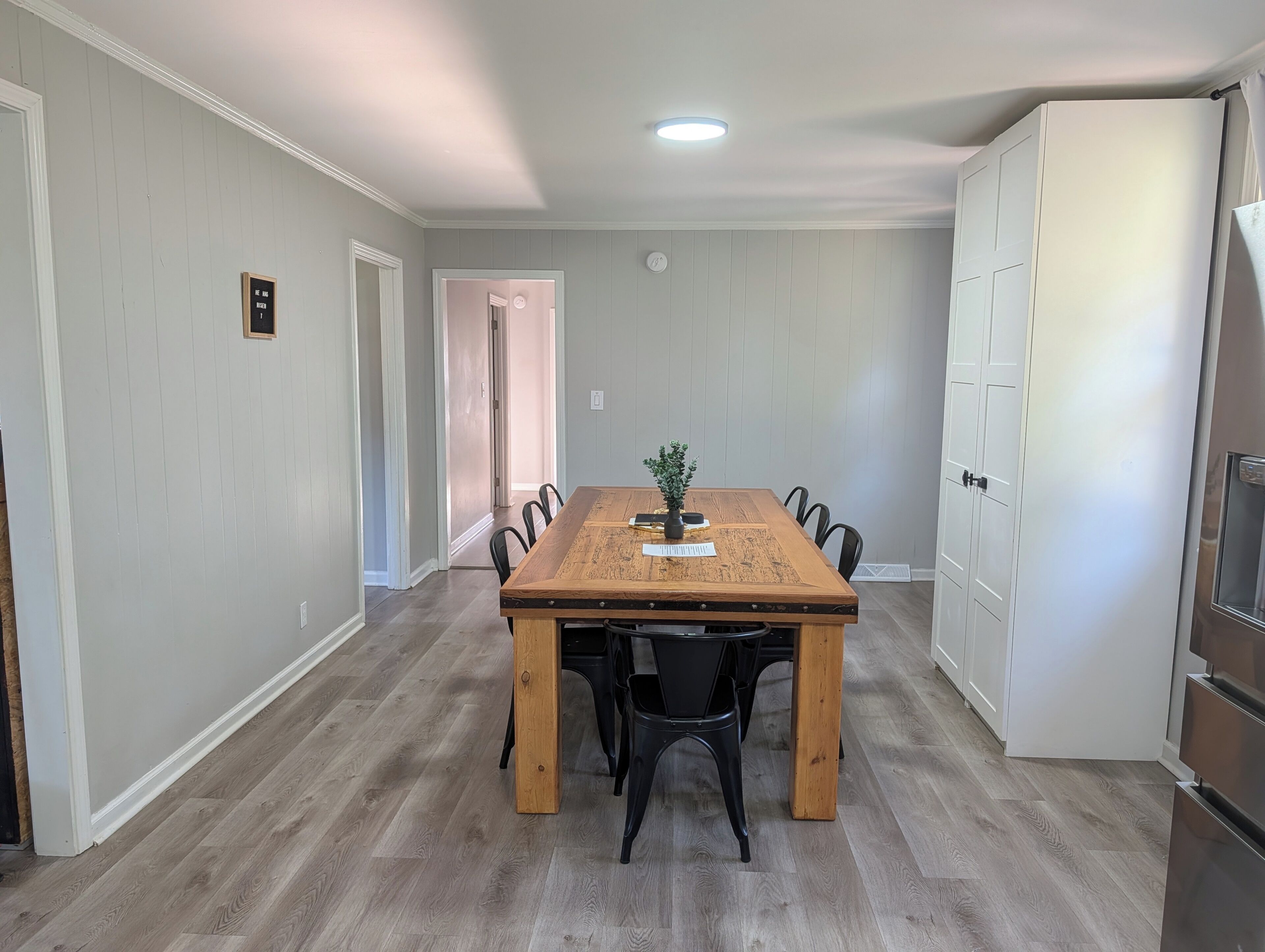 A dining area with a wooden table and black chairs, a small plant on the table, and a white door in the background.