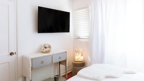 Un dormitorio con cama, mesita de noche con lámpara, televisor montado en la pared y ventana con persianas.