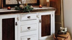 Un mueble blanco con cajones, un cuadro con una vaca, velas y un florero con flores.