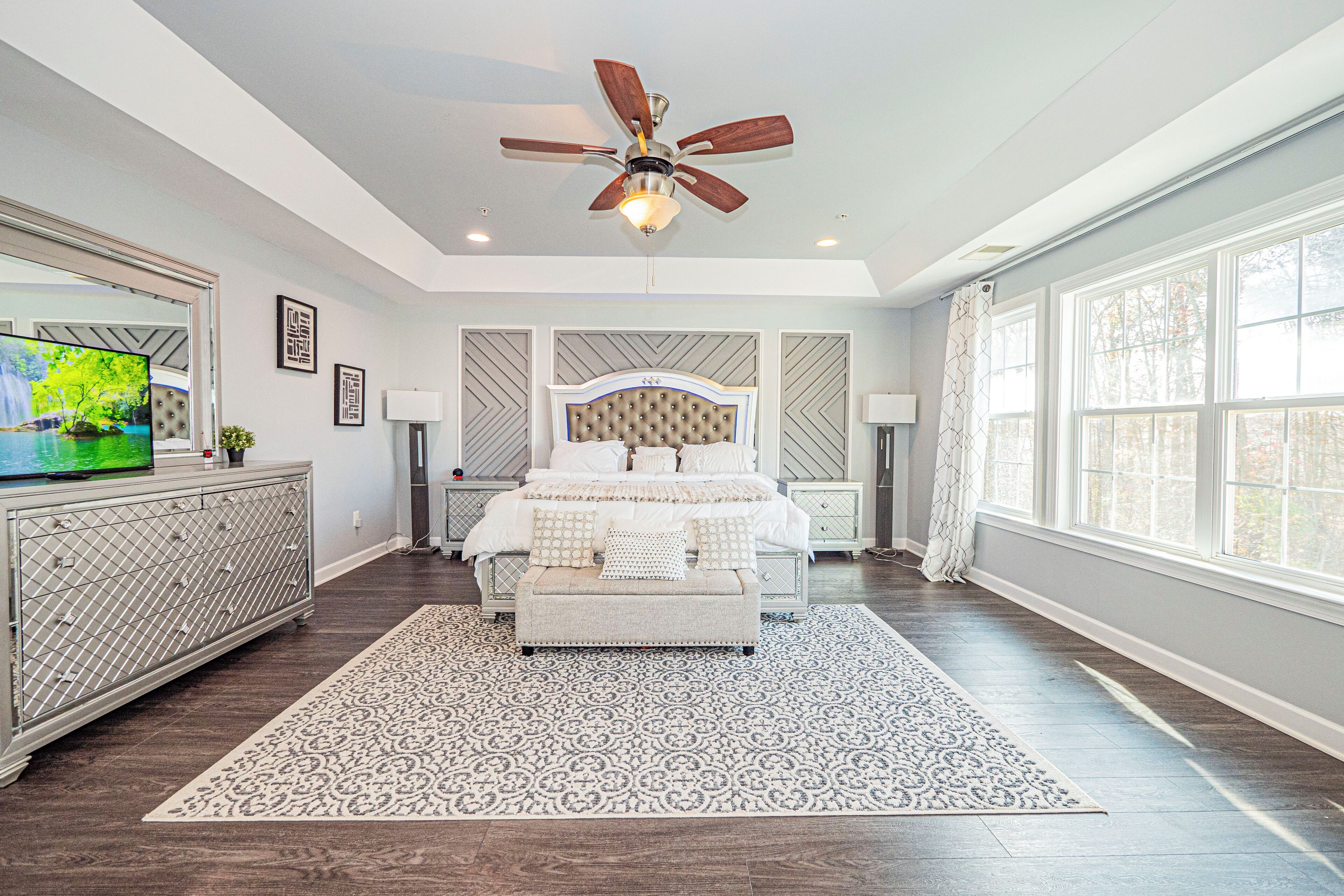 A bedroom with a large bed, a ceiling fan, a TV, and a window.