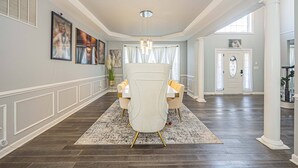 A spacious hallway with a dining area featuring a white chair and table, framed artwork on the walls, and a large window with curtains.