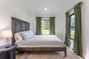 A bedroom with a bed, a bedside table, a window with curtains, and a view of greenery outside. - Modern Amish Country Retreat w/ Hot Tub & Spa Bath (Killbuck)
