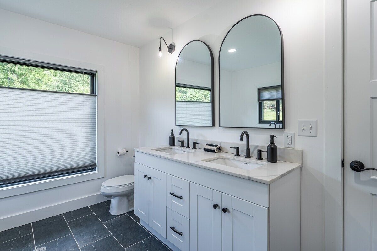 Een moderne badkamer met toilet, twee wasbakken, een spiegel en een raam.