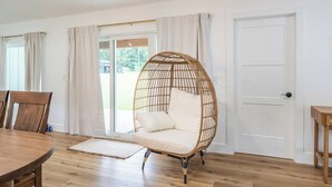 Ein Zimmer mit einem runden Holztisch, einem Korbstuhl mit Kissen, einer Holztür und Blick durch eine Glastür.