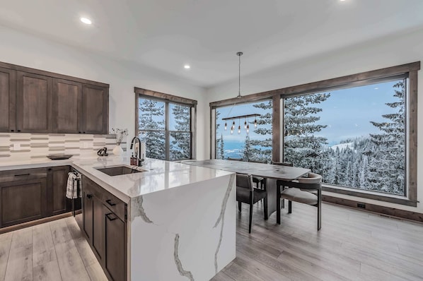 A modern kitchen with a large island, wooden cabinets, and a view of snow-covered trees through large windows.