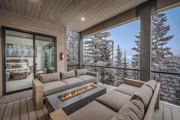 Un espacio exterior moderno con una fogata, asientos y vistas a árboles nevados.