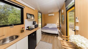 Een compacte kamer met een bed, een klein kitchenette, een raam met uitzicht op groen en een houten bankje met witte bloemen.