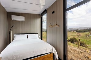 A bedroom with a large bed, a wall-mounted air conditioner, and a view of a rural landscape through a large window. - Melmarie Cosy Retreat X by Tiny Away (Waitati)