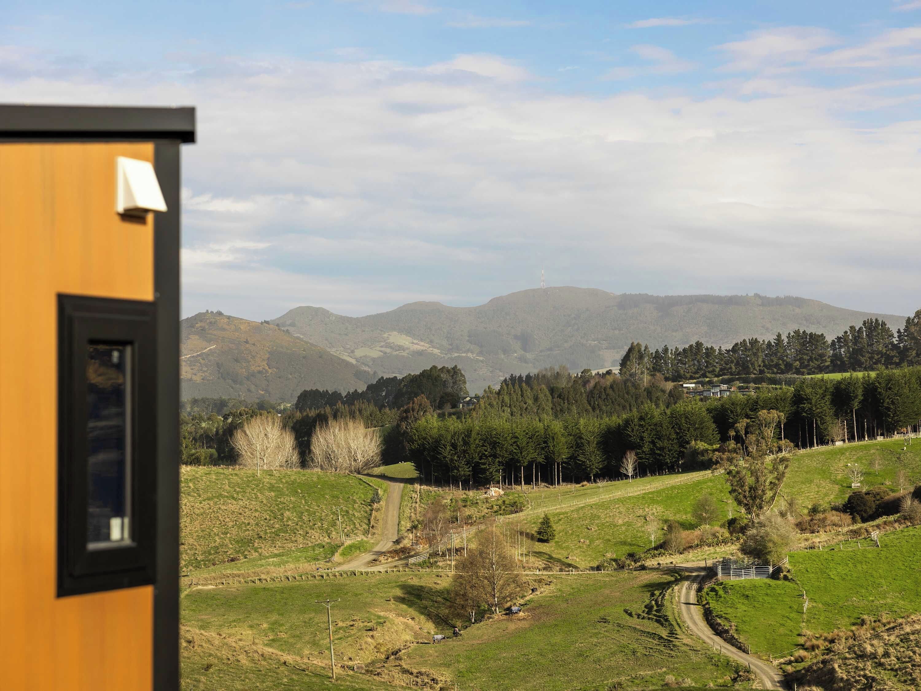 A view from a window of a building overlooking a rural landscape with rolling hills, a winding road, and a distant mountain range.