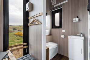 A modern bathroom with a toilet, sink, and mirror. There is a wooden bench and a view of a green landscape through a large window. - Melmarie Cosy Retreat X by Tiny Away (Waitati)