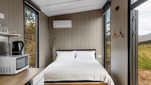A modern bedroom with a wooden bed, a white bedside table with a microwave, and a view of a natural landscape through large windows. - Melmarie Cosy Retreat X by Tiny Away (Waitati)