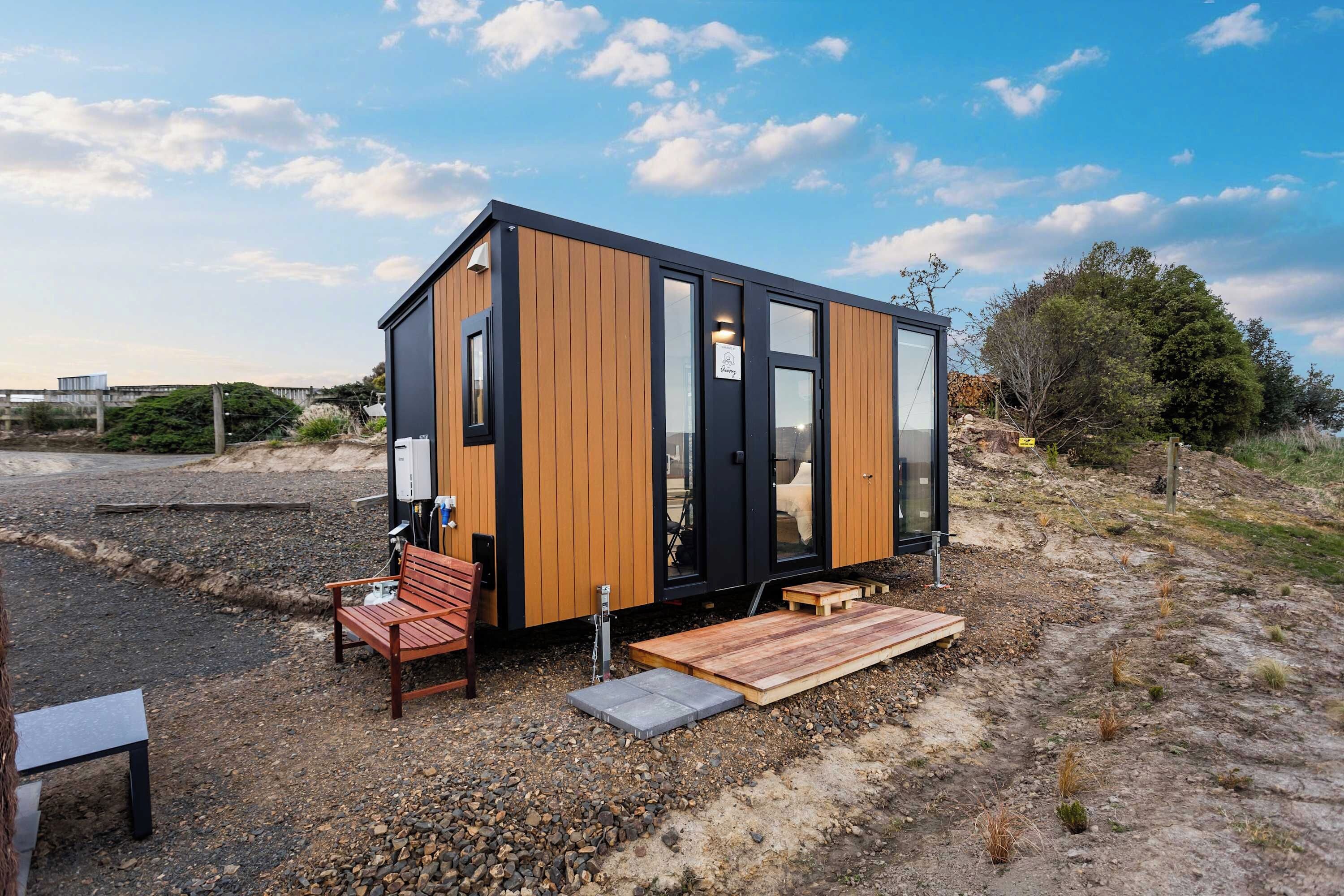 Uma cabana moderna e compacta com exterior de madeira e janelas grandes, com um banco e uma mesa pequena na parte externa.