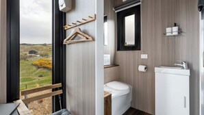 Ein modernes Badezimmer mit Toilette, Waschbecken und Spiegel. Eine Holzbank und ein Blick auf eine grüne Landschaft durch ein großes Fenster.