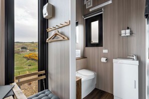 A modern bathroom with a toilet, sink, and mirror. There is a wooden bench and a view of a green landscape through a large window. - Melmarie Cosy Retreat X by Tiny Away (Waitati)