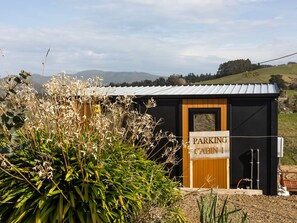 Une petite cabane avec une pancarte indiquant « CABINE DE STATIONNEMENT » devant un bâtiment à toit métallique, entourée de verdure et avec des collines en arrière-plan.