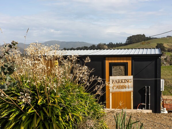 Una cabaña pequeña con un letrero que dice "PARKING CABIN" frente a un edificio con techo de metal, rodeada de follaje y colinas al fondo.