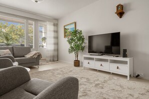 A living room with a large flat-screen TV, a white entertainment unit, a grey sofa, and a large window with blinds.
