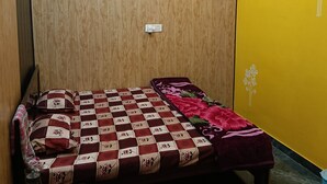A room with a wooden wardrobe, a bed with a floral quilt, a small table with glasses, and a yellow wall with a floral design.