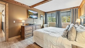 A bedroom with a large bed, wooden beams, a dresser, a TV, and a window with curtains. - Breck Condo | Private Balcony & Trail Access (Breckenridge)