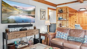 A cozy living room with a large TV, wooden furniture, and a leather couch. - Breck Condo | Private Balcony & Trail Access (Breckenridge)