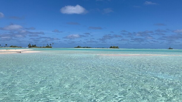 On peut admirer une plage avec une eau turquoise, un rivage sablonneux et des îles lointaines, le tout sous un ciel bleu parsemé de nuages.